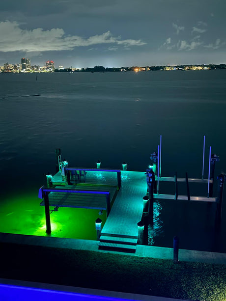 Dock illuminated with green and blue piling cap lights at night over a body of water with city skyline in the background.